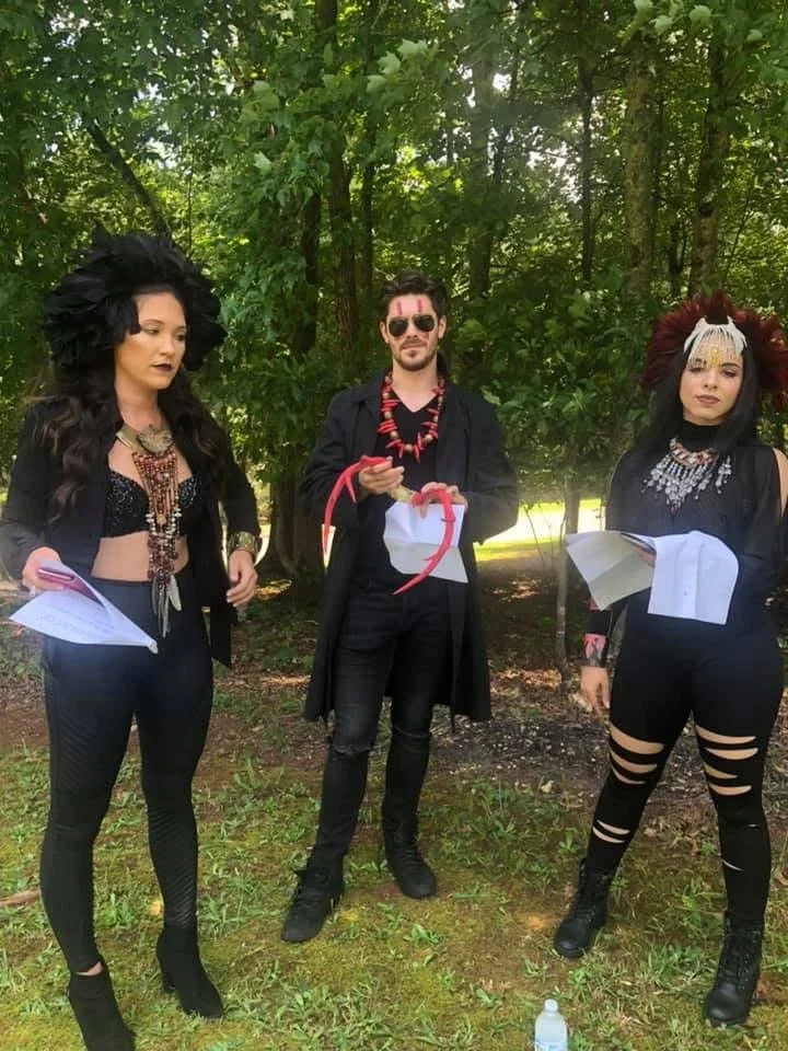 Three people dressed in dark, gothic clothing with elaborate makeup and accessories stand outdoors among trees, holding papers and props.