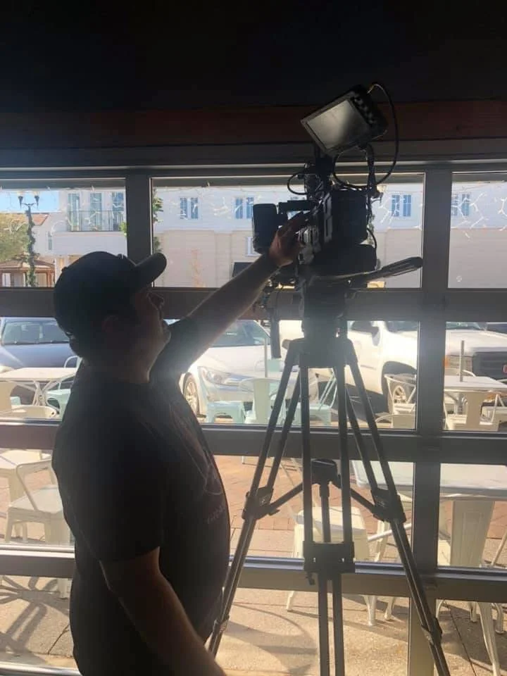 Person operating a video camera on a tripod inside a room with large windows, overlooking outdoor seating and parked cars.