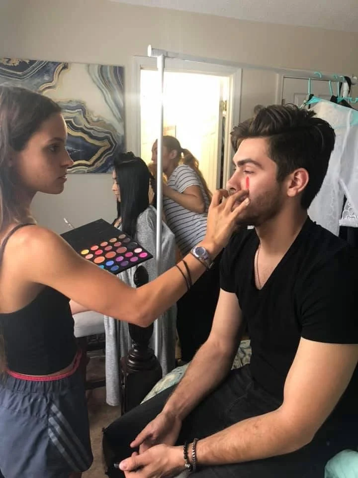 A woman applying makeup to a man, who is sitting in a room with two other women in the background. The woman is holding a makeup palette, and the man's face is being touched up with makeup.