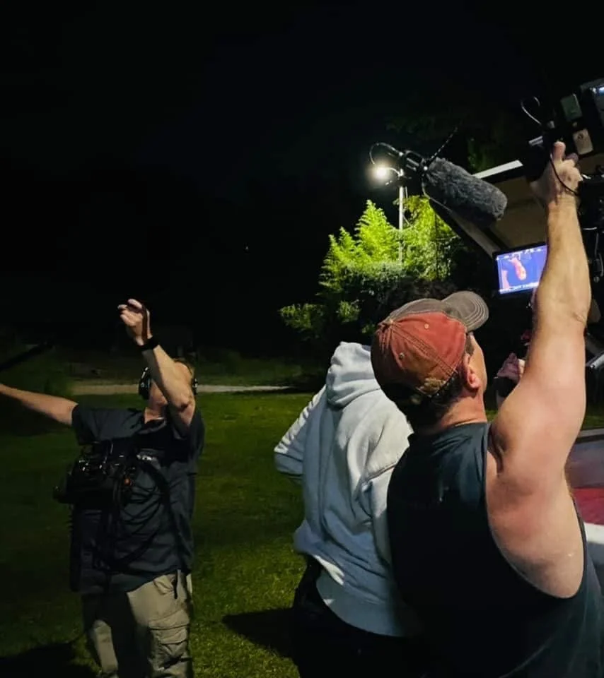 Nighttime filming scene outdoors with three crew members; one man is holding a camera, another is adjusting a light overhead, and a third is framing a shot in the distance.