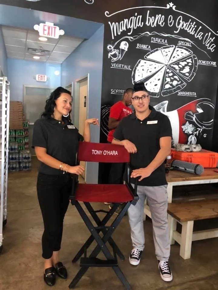 Two people standing inside a restaurant or cafe, with a woman on the left and a man on the right, both wearing black shirts. They are posing next to a director's chair mounted on a stand, which has the name 'Tony O'Hara' on the backrest. Behind them,