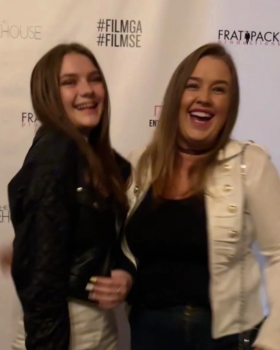Two women smiling and posing together at an event with a backdrop featuring logos and hashtags.