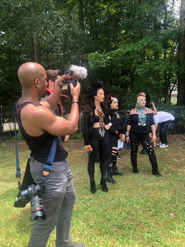 Photographer taking a picture of four women dressed in edgy, goth-inspired clothing in a wooded outdoor setting.