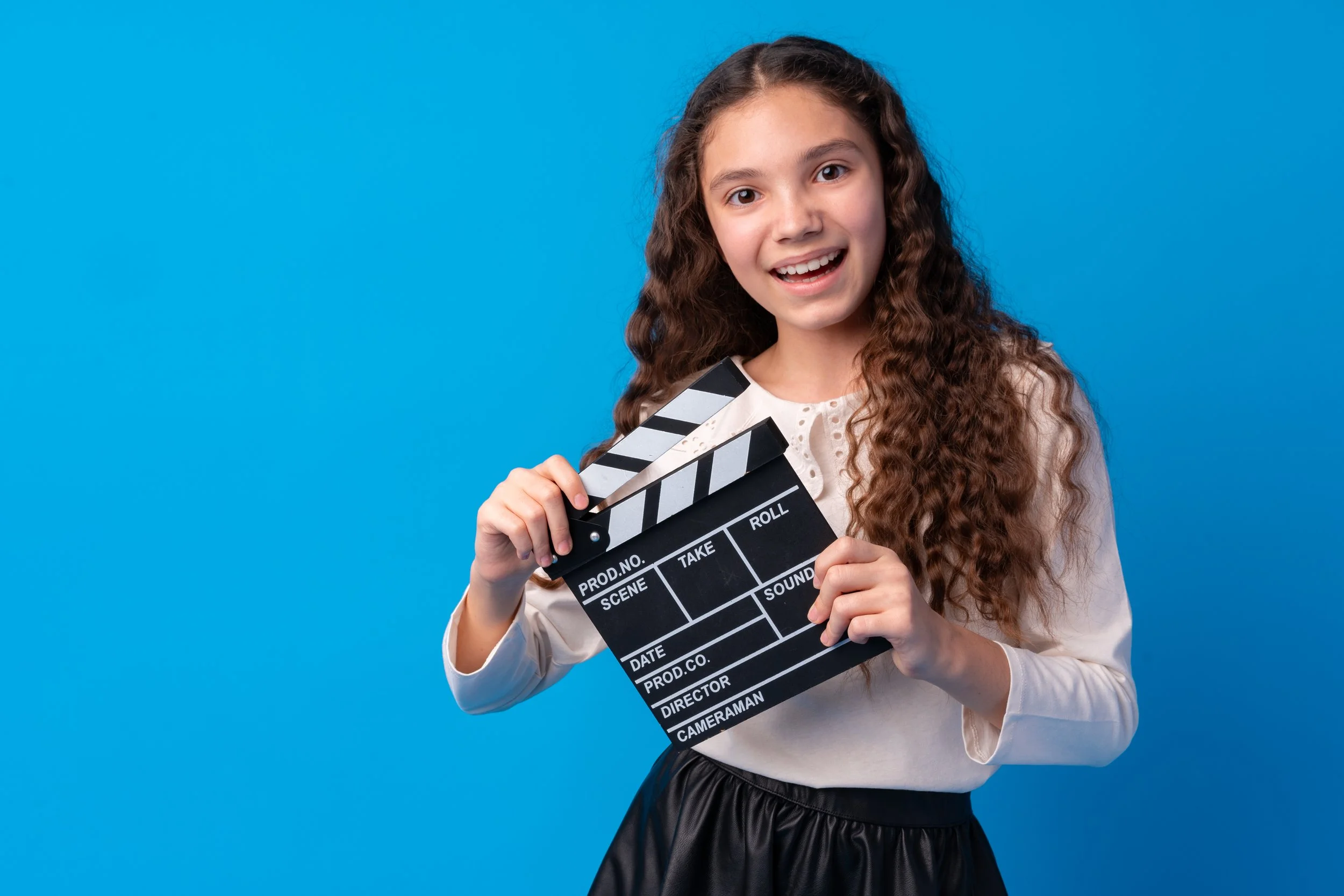 Young girl with long curly hair holding a film clapperboard and smiling against a bright blue background.