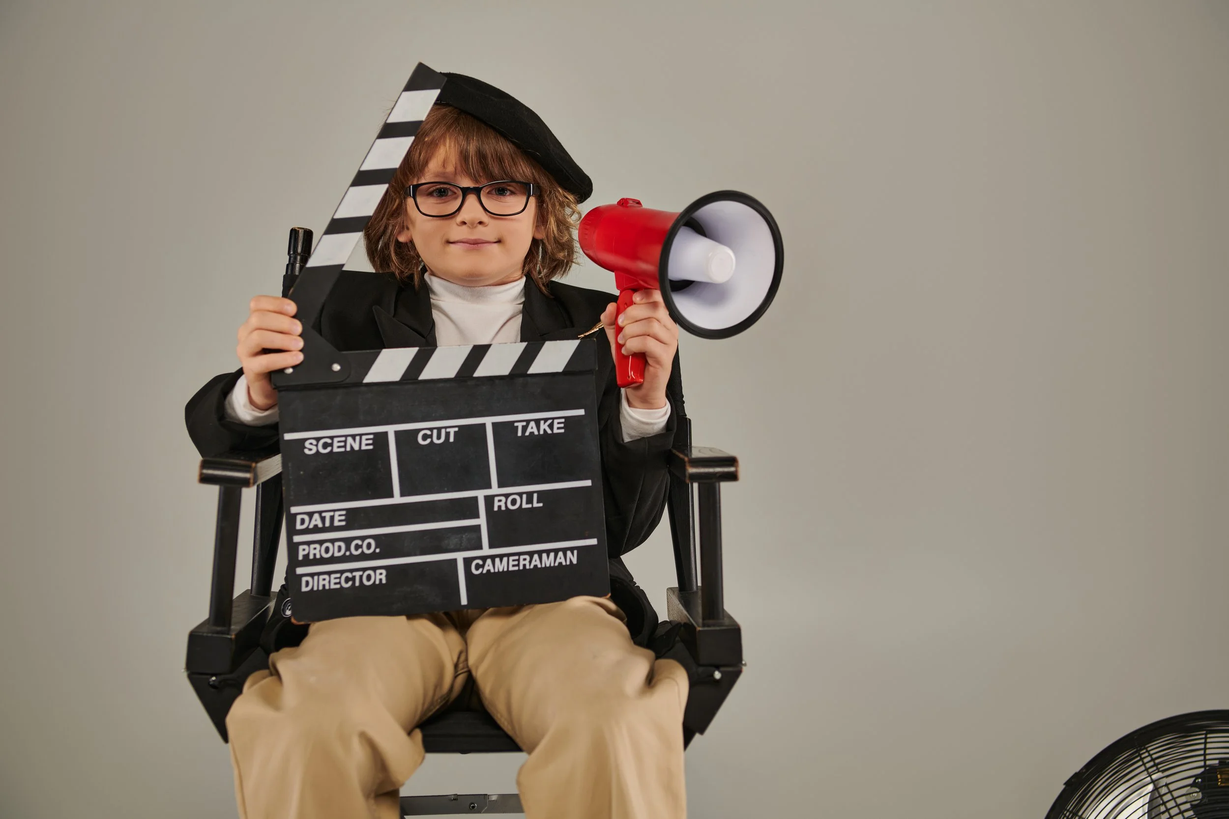A young child sitting in a director's chair, wearing a black hat and glasses, holding a clapperboard in one hand and a red megaphone in the other, against a plain light-colored background.