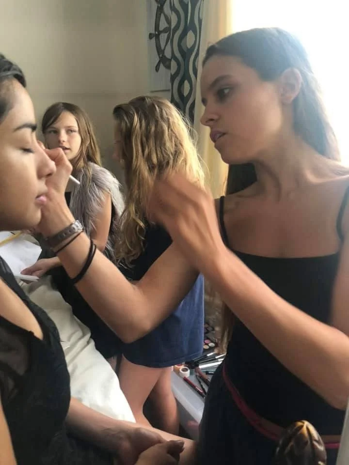 Makeup artist applying makeup to a young woman's face surrounded by young girls in a room with patterned curtains.