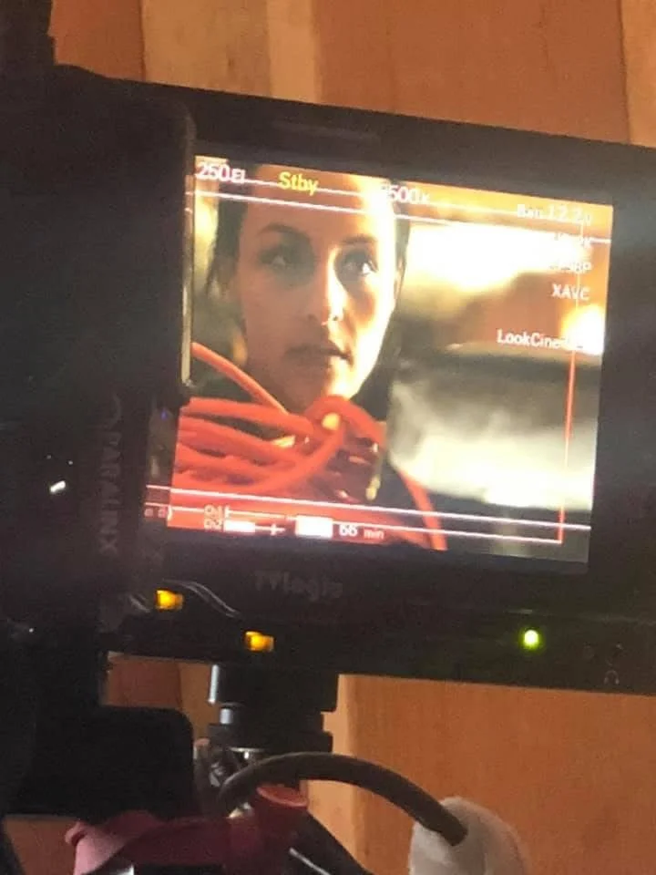 Photograph of a camera monitor displaying a close-up of a woman with dark hair and eyes, wearing a red scarf, in front of a wooden background.