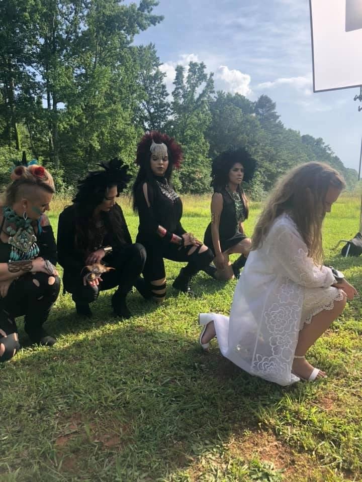 A woman dressed in a white lace dress kneeling in front of four women dressed in punk and gothic style outfits outdoors on a grassy field with trees in the background, with a clear sky and some clouds.