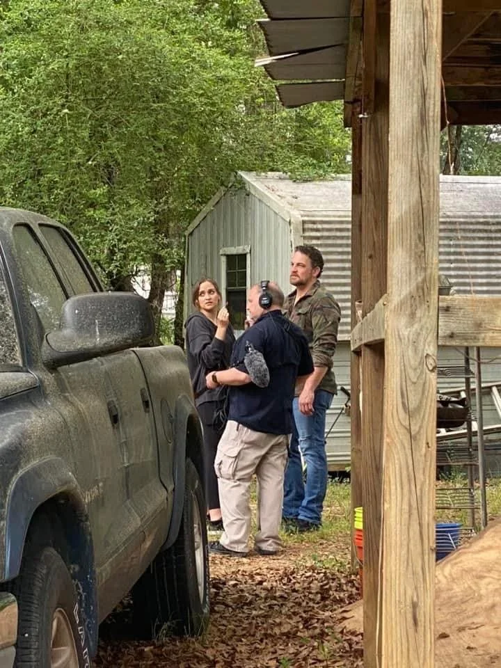 Three people are outdoors, two men and one woman, engaged in conversation near an old building. One man wears headphones and is holding a microphone. The woman looks thoughtful. A muddy pickup truck is in the foreground, and a small shed with trees i