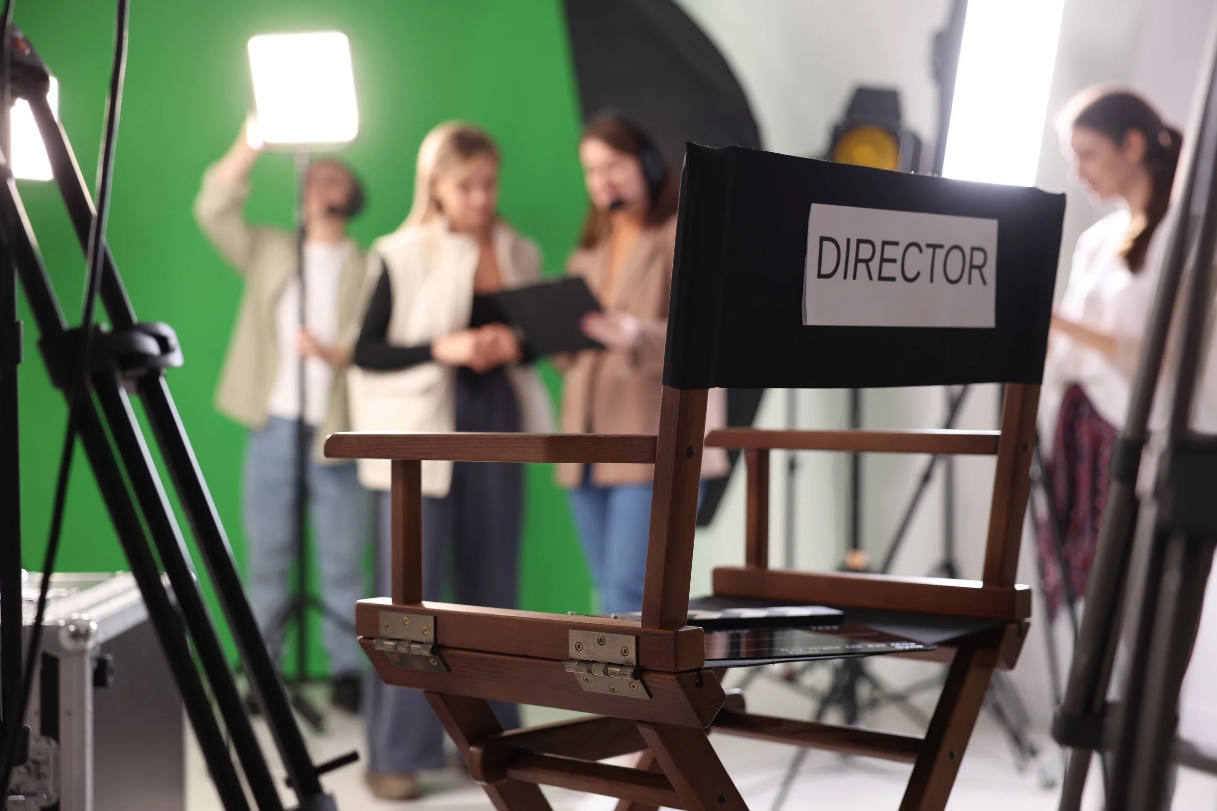 A director's chair with a white sign that says 'DIRECTOR' in a film studio with a green screen and crew members in the background.
