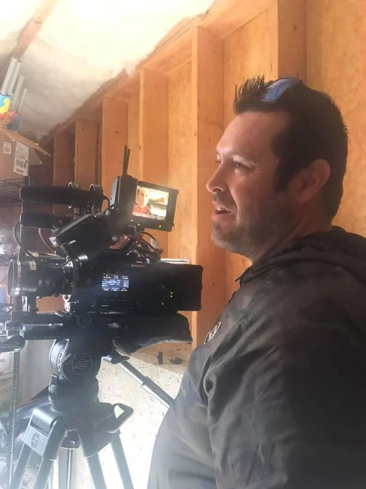 A man operating a professional video camera inside a wooden structure, possibly a garage or workshop, with wood framing visible in the background.