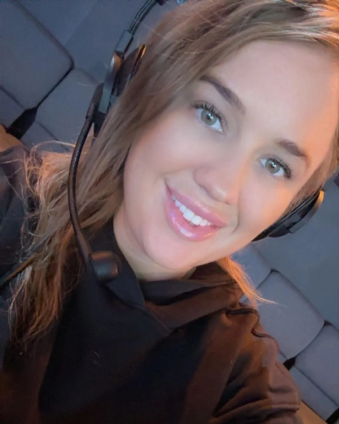 A woman with light brown hair smiling, wearing a black top, headphones, and a jacket, sitting on gray seats.