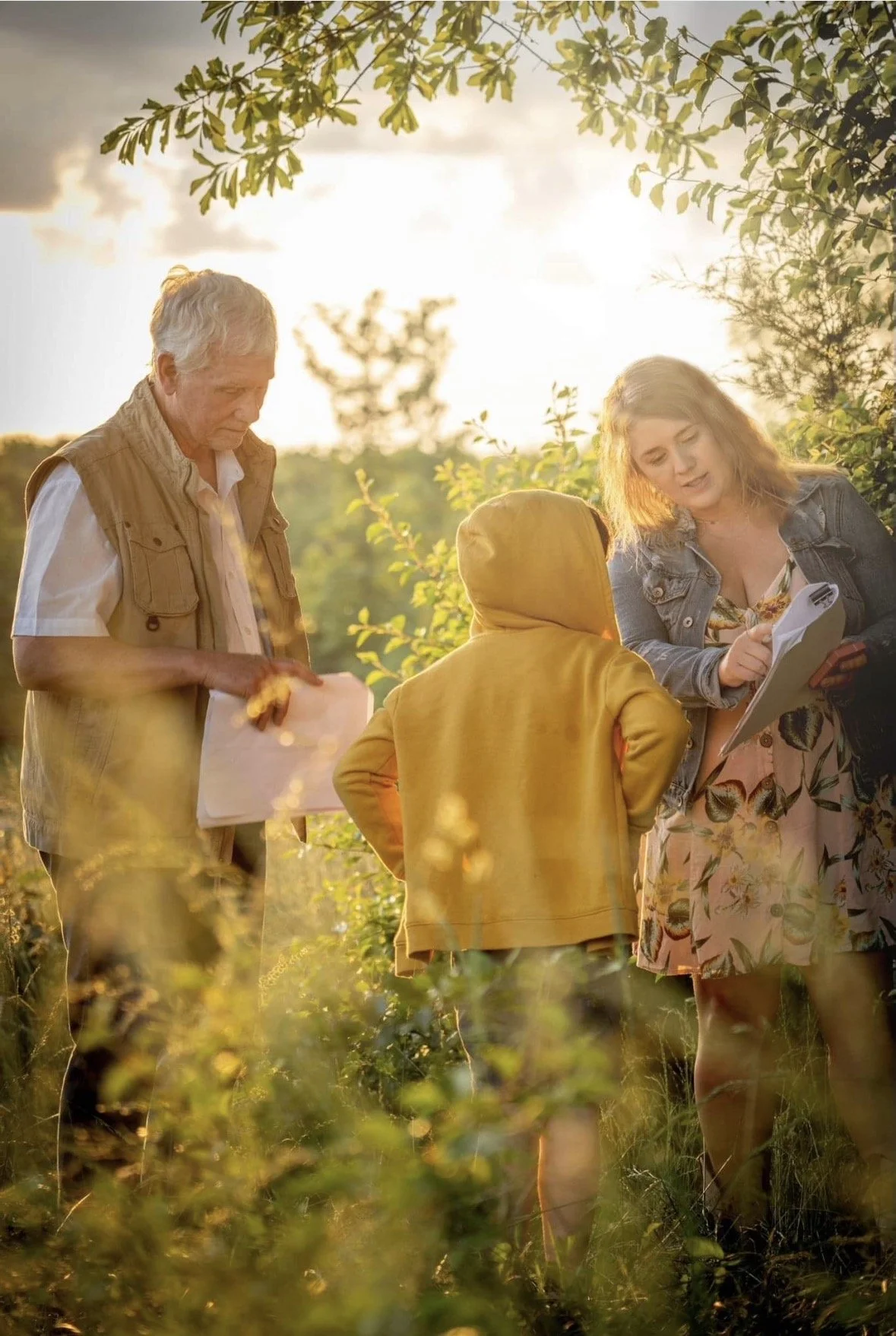 Three people, two women and one young boy, are outdoors in a sunny field, looking at a piece of paper or a map, with trees and sunlight in the background.