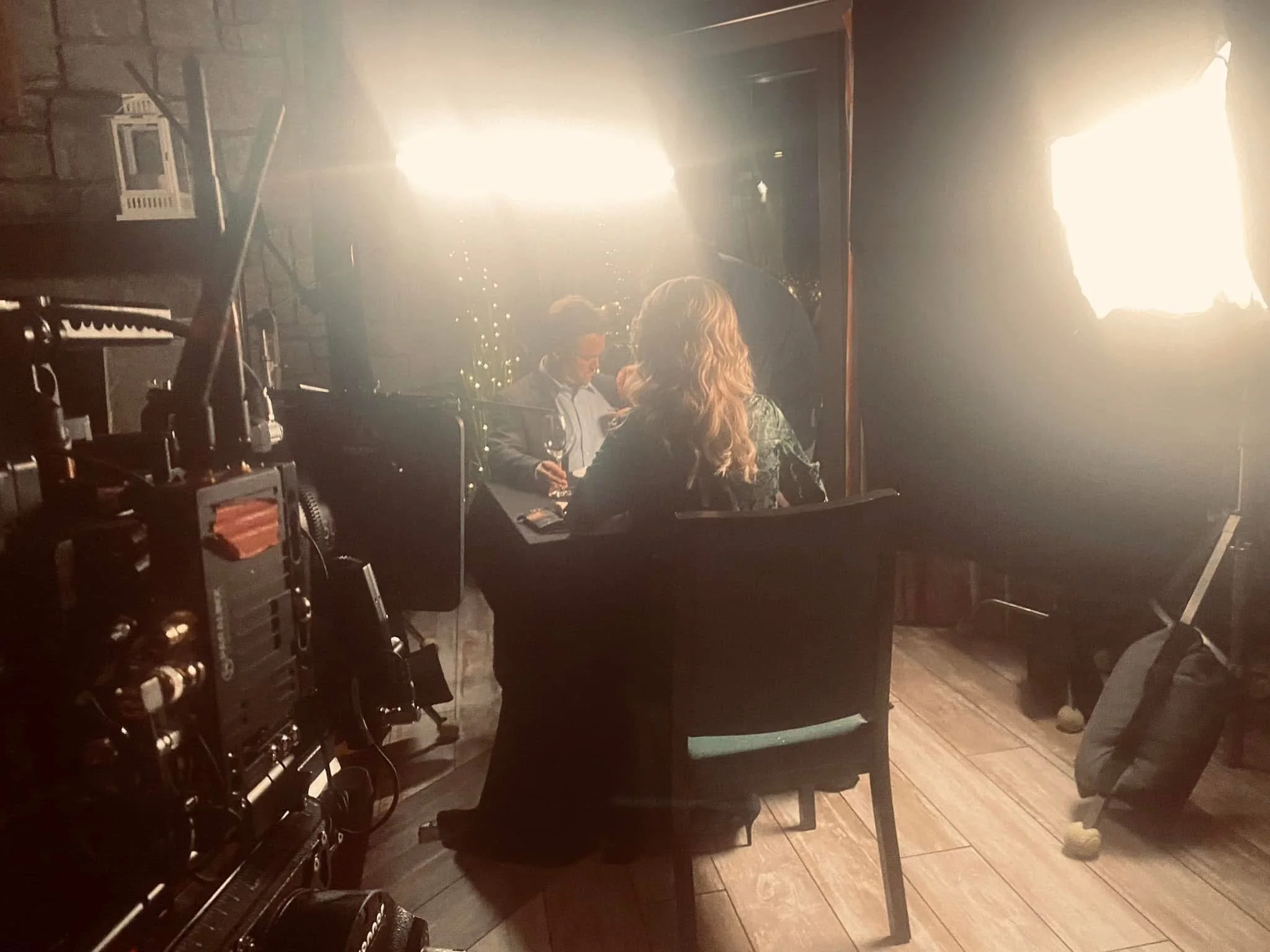 A dimly lit room with a large bright light source creating a glow. A woman with blonde hair and a man in a blazer are sitting at a table, engaged in conversation, with the woman holding a glass of wine. There is a brick wall in the background, and eq