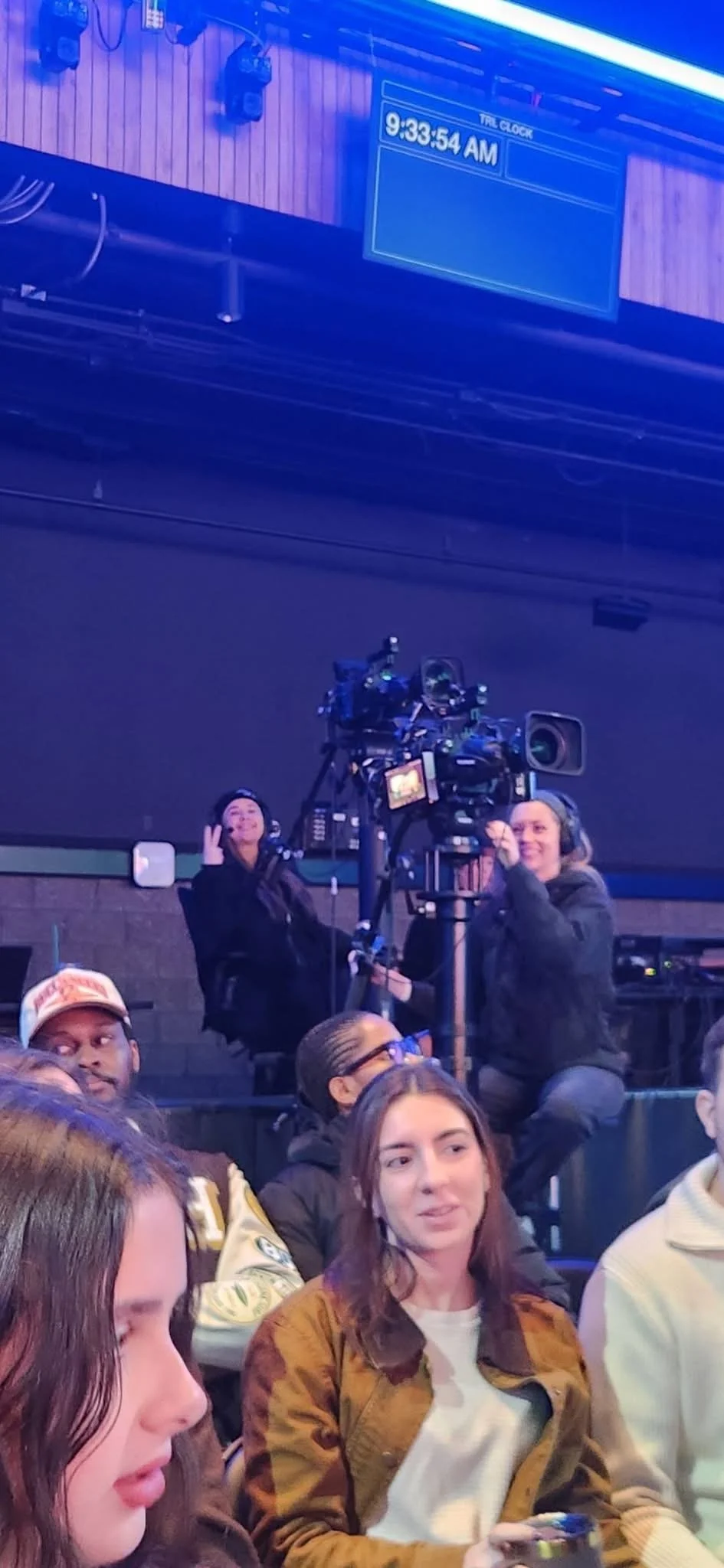 Scene inside a hospital or waiting area with people sitting and a camera setup. Two women are operating a professional camera on a tripod, one adjusting the camera and the other smiling. Several other individuals are visible, some looking at the came