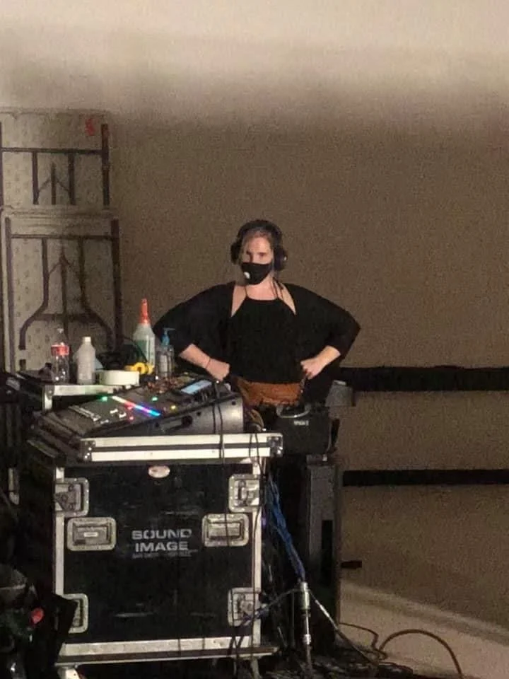 A woman DJ wearing headphones and a face mask stands behind DJ equipment in a dimly lit room.