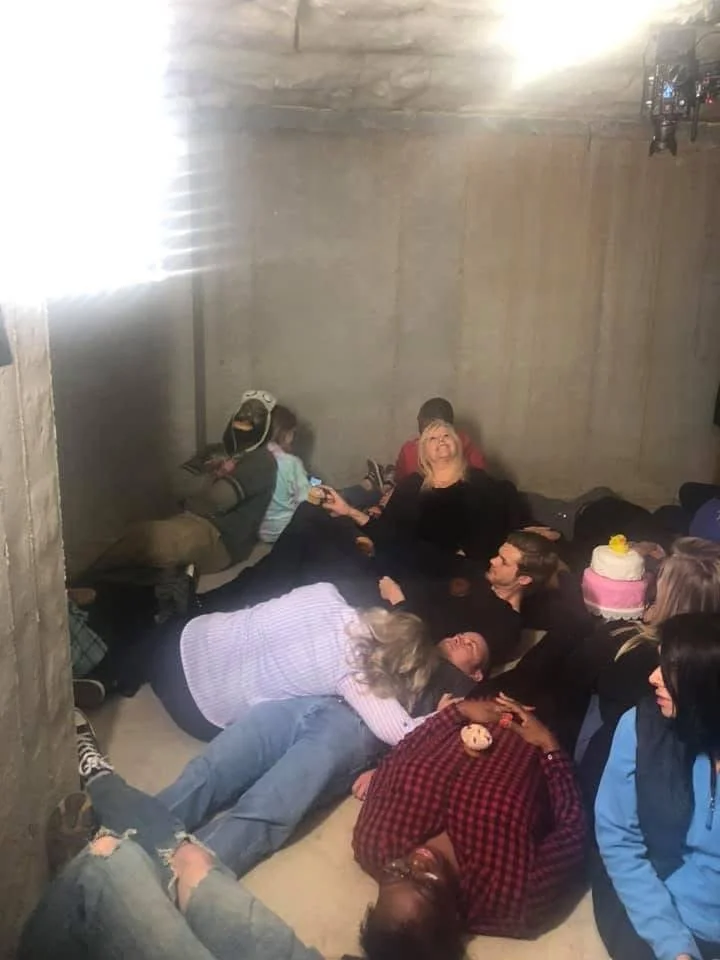 Several people are lying on the floor in a small, concrete room, some laughing and others resting, with a woman smiling and looking up in the center of the group.