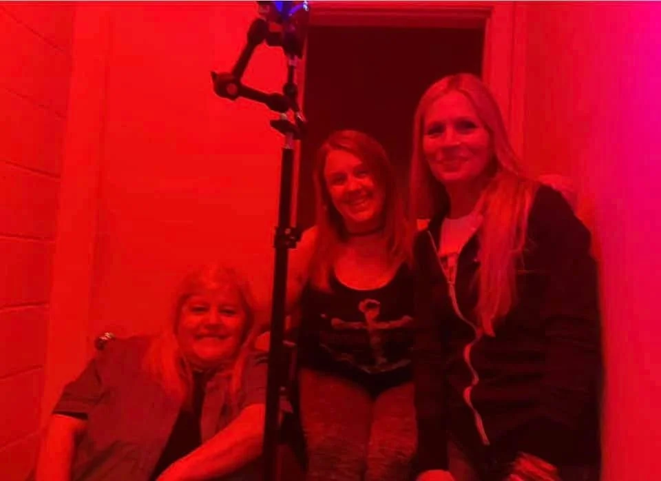 Three women smiling inside a room bathed in red light, one seated and two standing, with a microphone stand in the foreground.