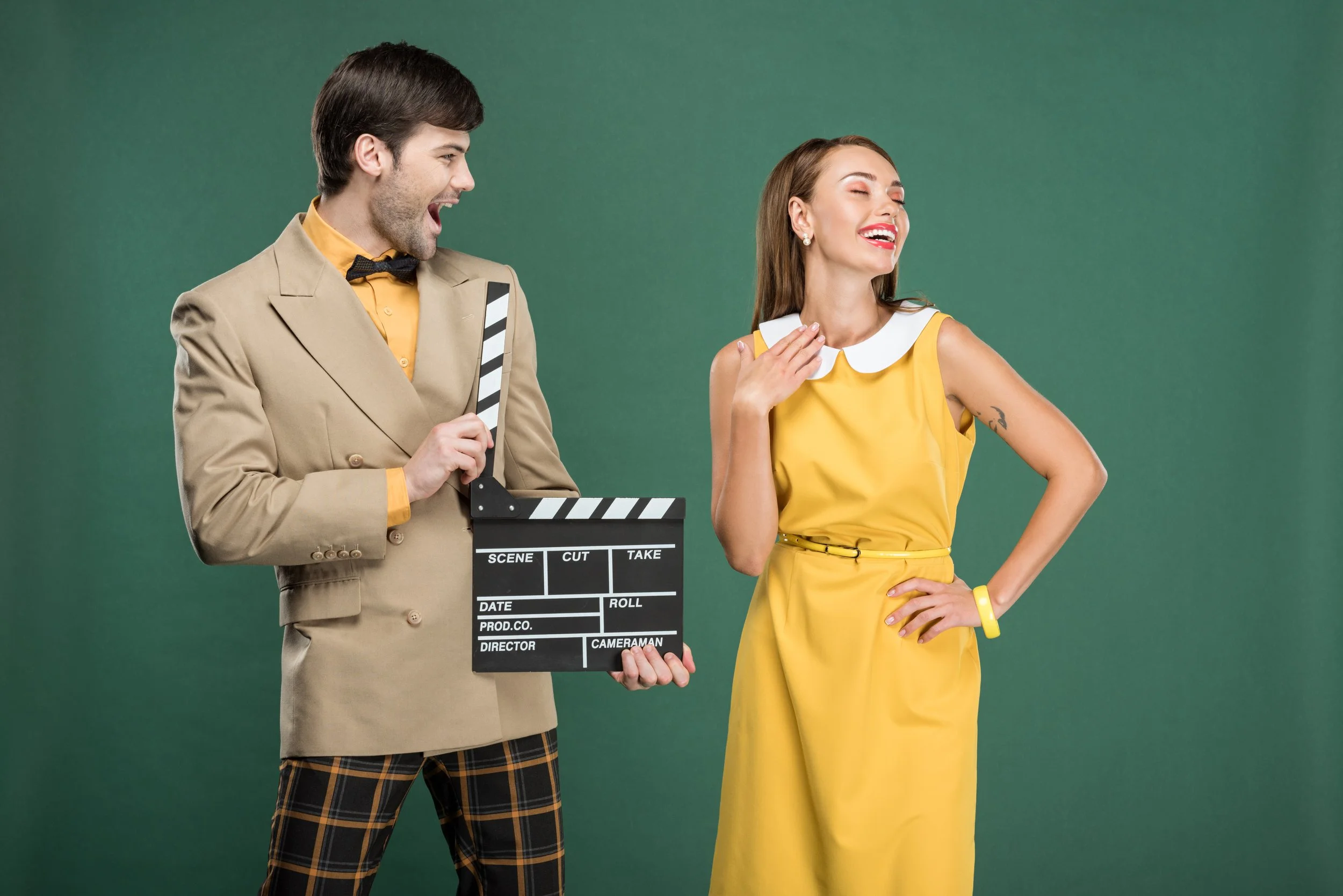 A man wearing a beige suit, yellow shirt, and plaid pants holding a clapperboard and laughing while a woman in a yellow dress with a white collar and a tattoo on her arm is laughing with eyes closed, hand on her chest, against a green background.