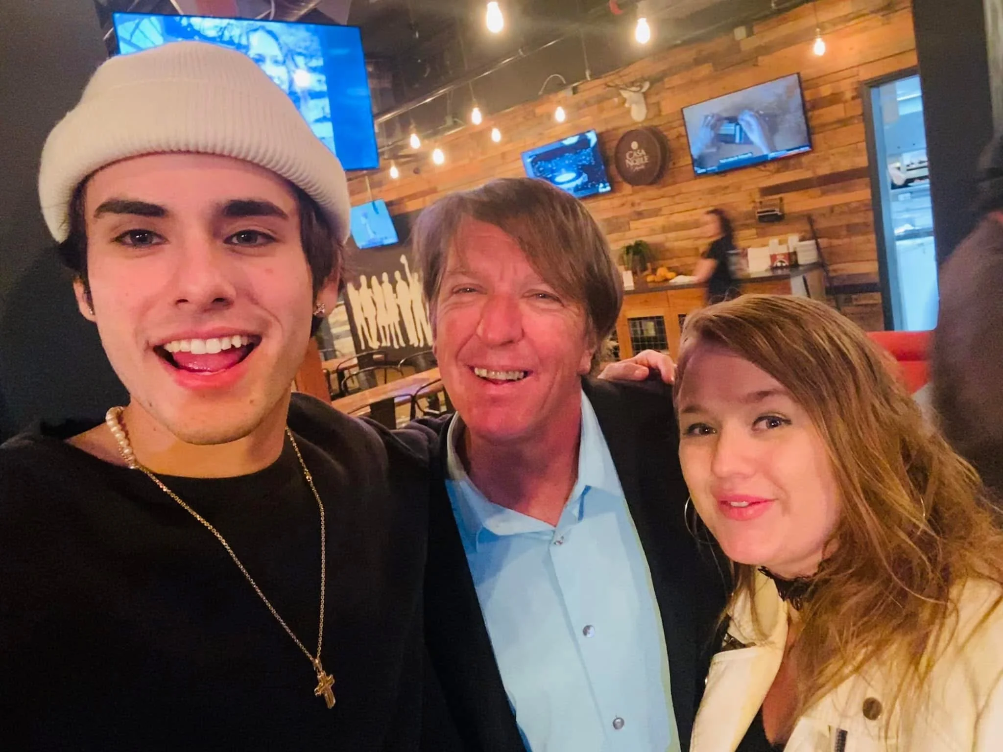 Young man with a white beanie, earrings, and pearl necklace, smiling with his tongue slightly out, standing next to older man and woman inside a restaurant or bar with wooden walls and hanging light bulbs.