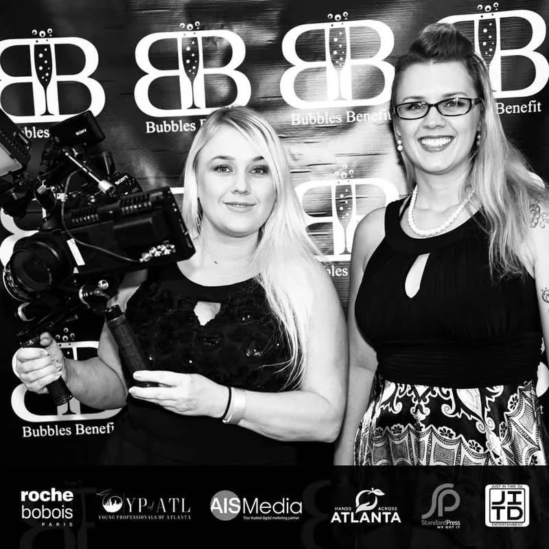 Two women posing for a photo at a Bubbles Benefit event, with a camera in hand and standing in front of a step and repeat banner.