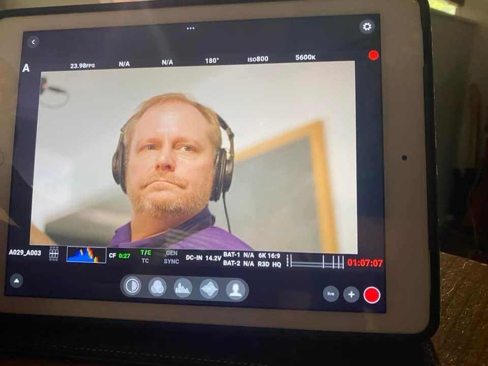 A man with blonde hair and facial hair wearing headphones is being recorded on a digital camera or tablet, with a blurred background that includes a chalkboard.