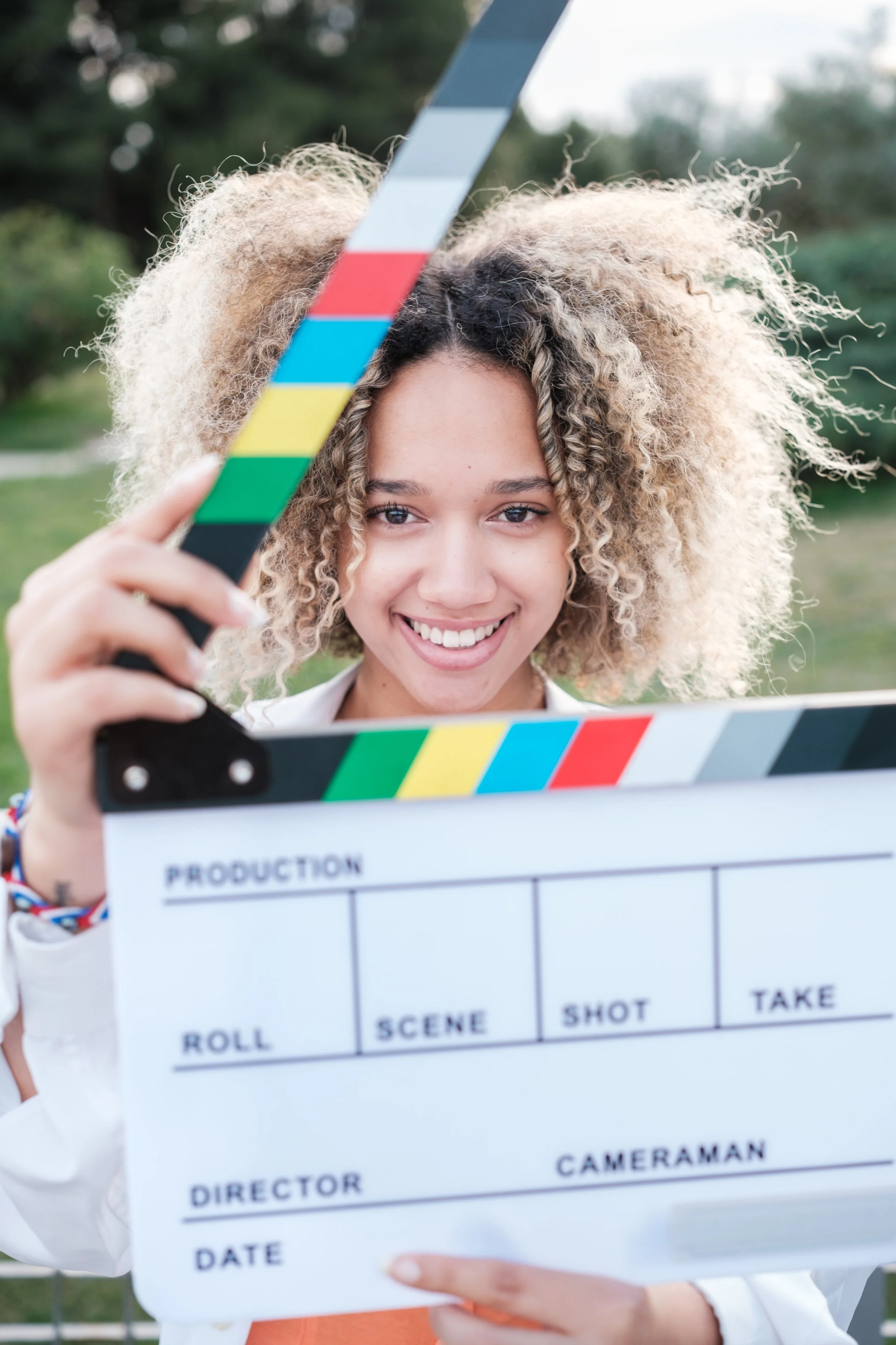 A woman is holding a film slate with a smile on her face, outdoors with green trees in the background.