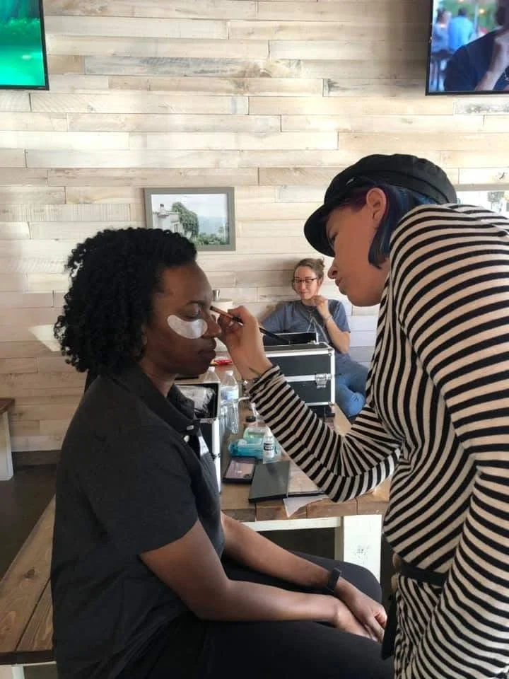 A woman with dark curly hair is having her face and eyes makeup applied by a makeup artist with short hair, wearing a striped long sleeve shirt.