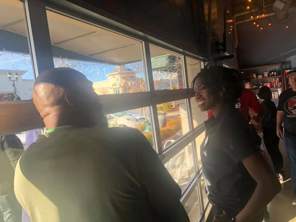 Two women are talking and smiling inside a café or restaurant, with large windows and outdoor décor visible behind them.