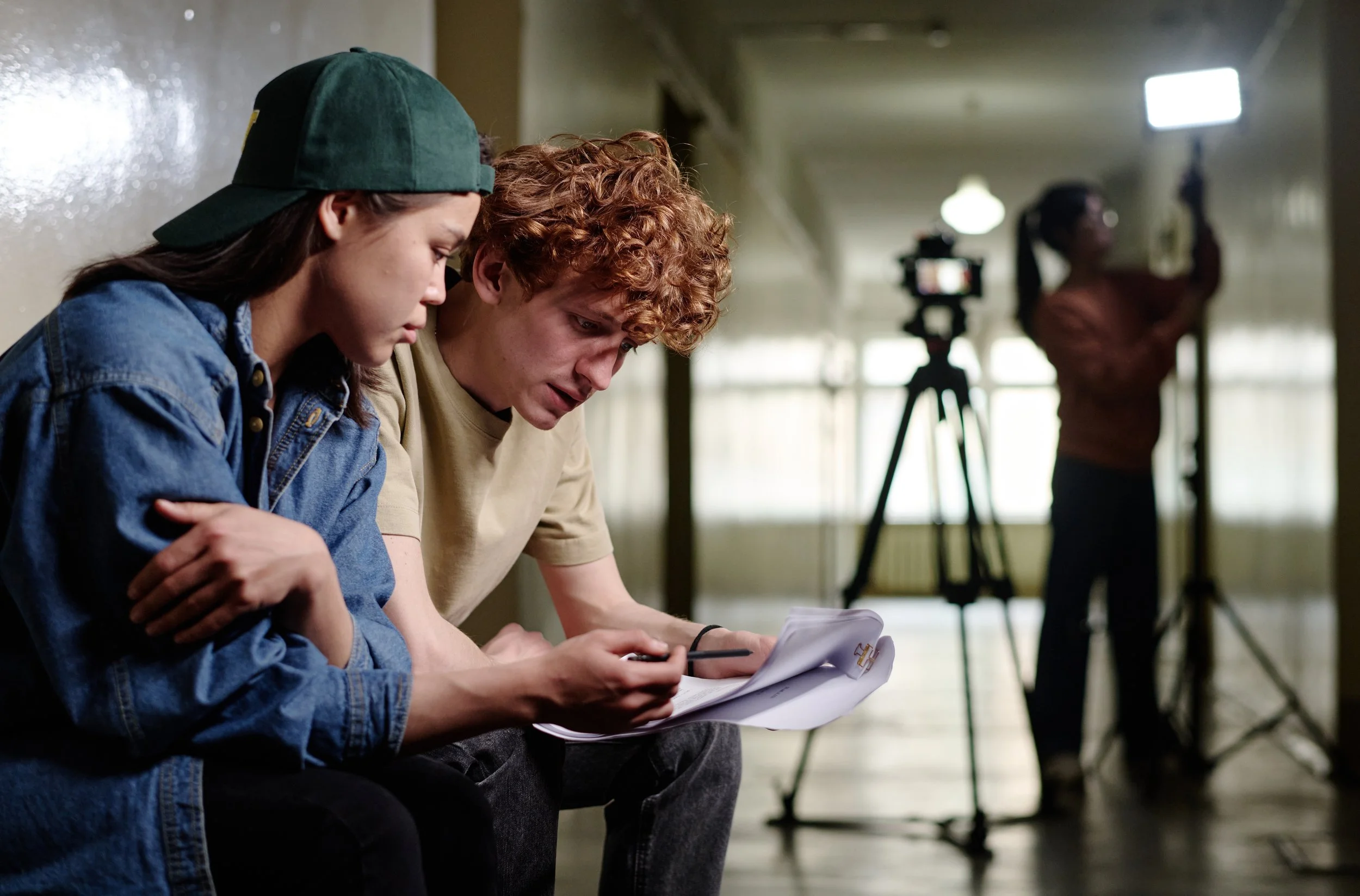 Two young people sit on a bench, closely reading a sheet of paper or a booklet. Behind them, a woman stands near a camera on a tripod, adjusting or inspecting her phone, in a hallway with large windows and light fixtures.