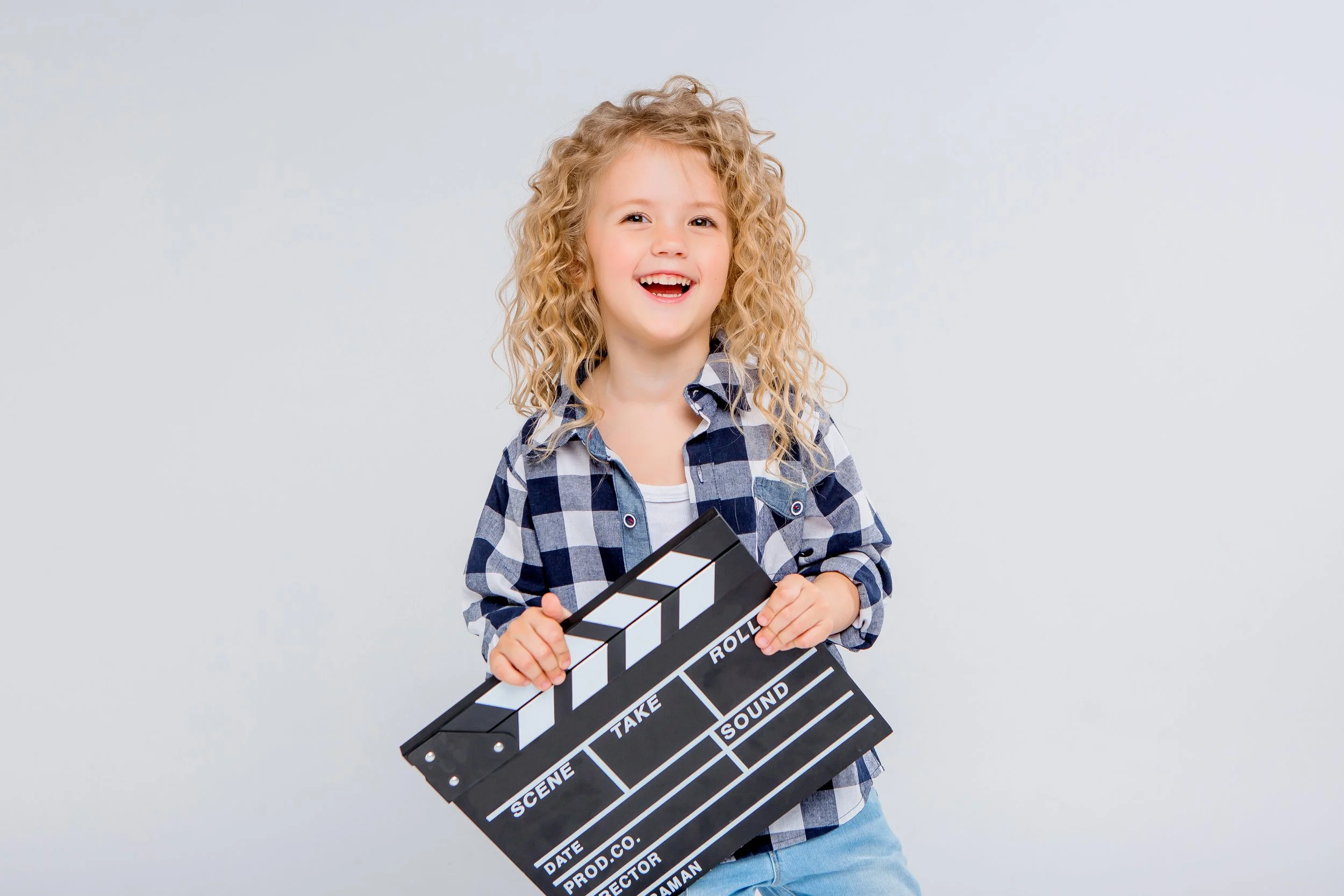 A young girl with curly blonde hair smiling and holding a black and white film slate.