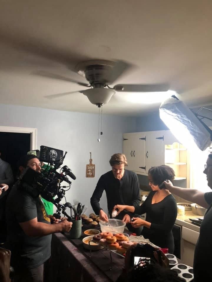 Filming crew recording a kitchen scene with two women preparing donuts while a man assists, in a well-lit kitchen with modern cabinets.