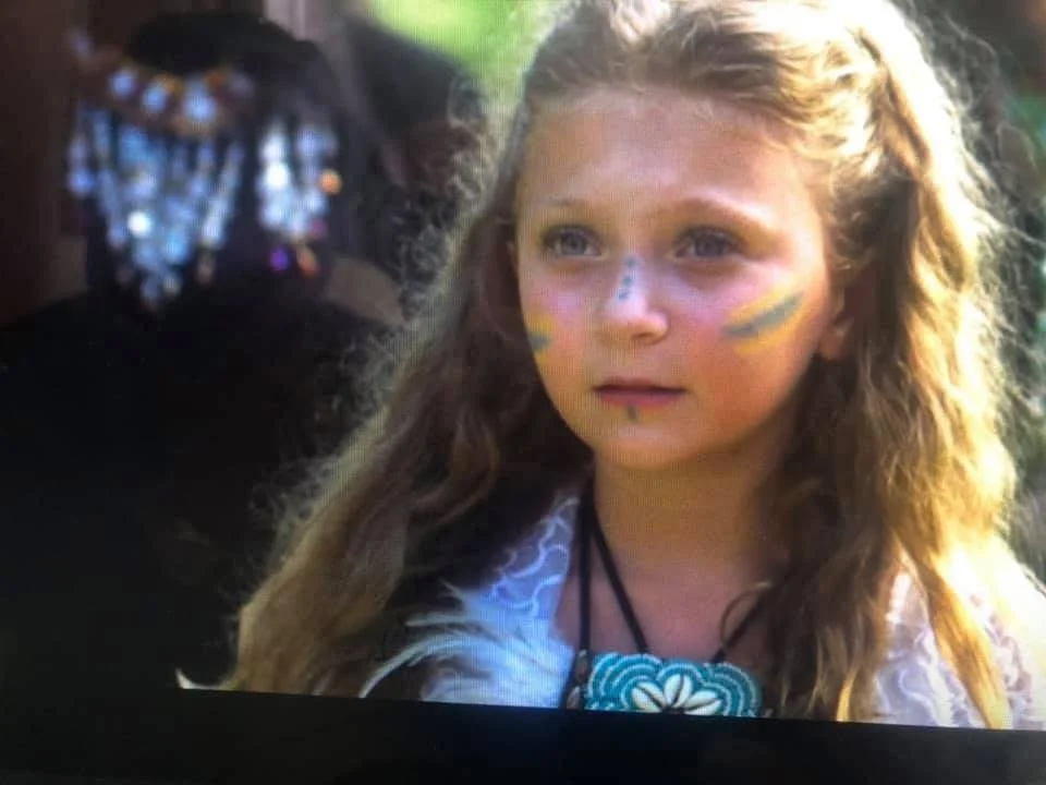 A young girl with long, curly hair and face paint, looking thoughtfully into the distance, outdoors with a blurred natural background.