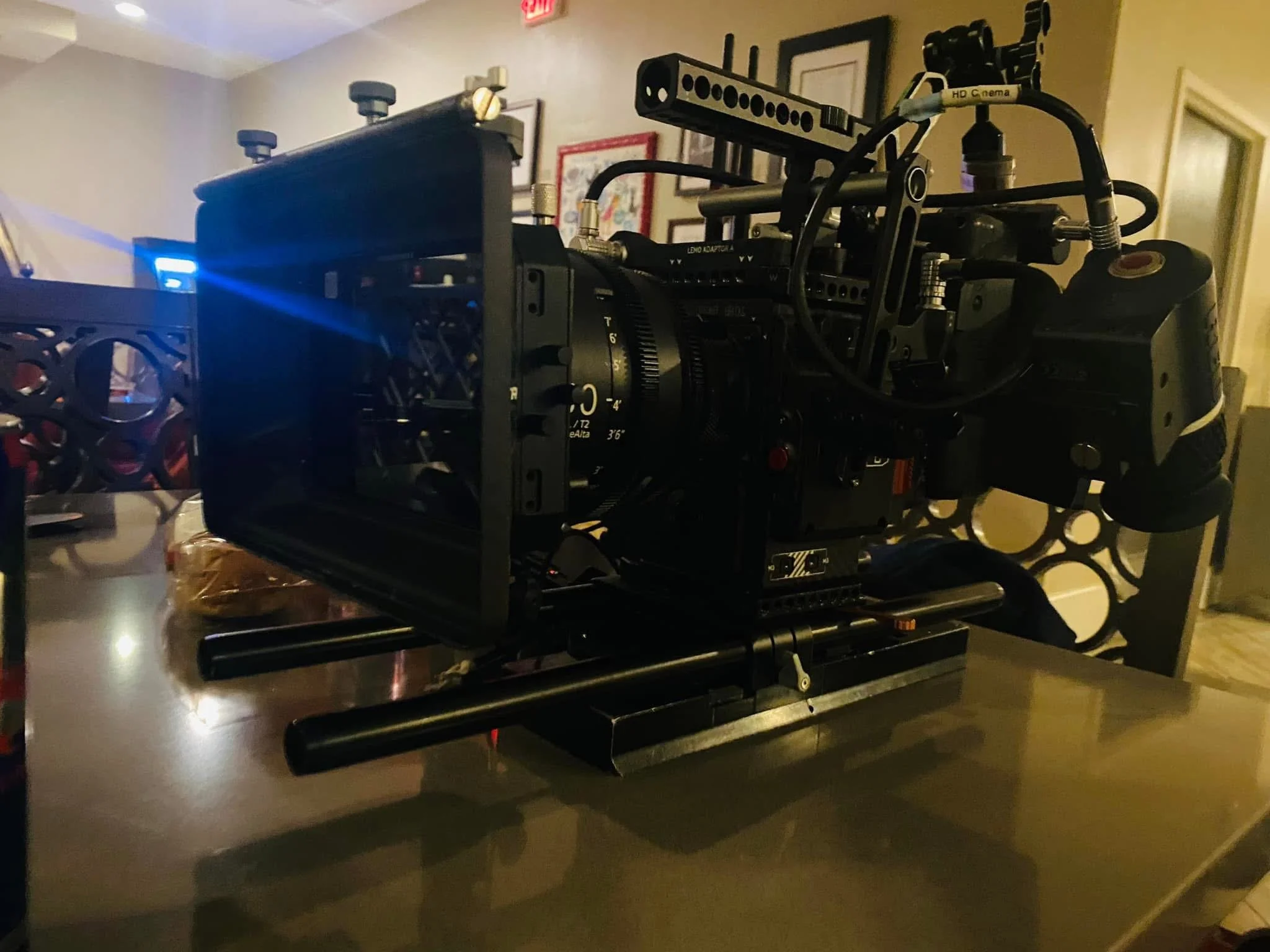 A professional cinema camera on a dolly, set up in a room with framed pictures on the wall.