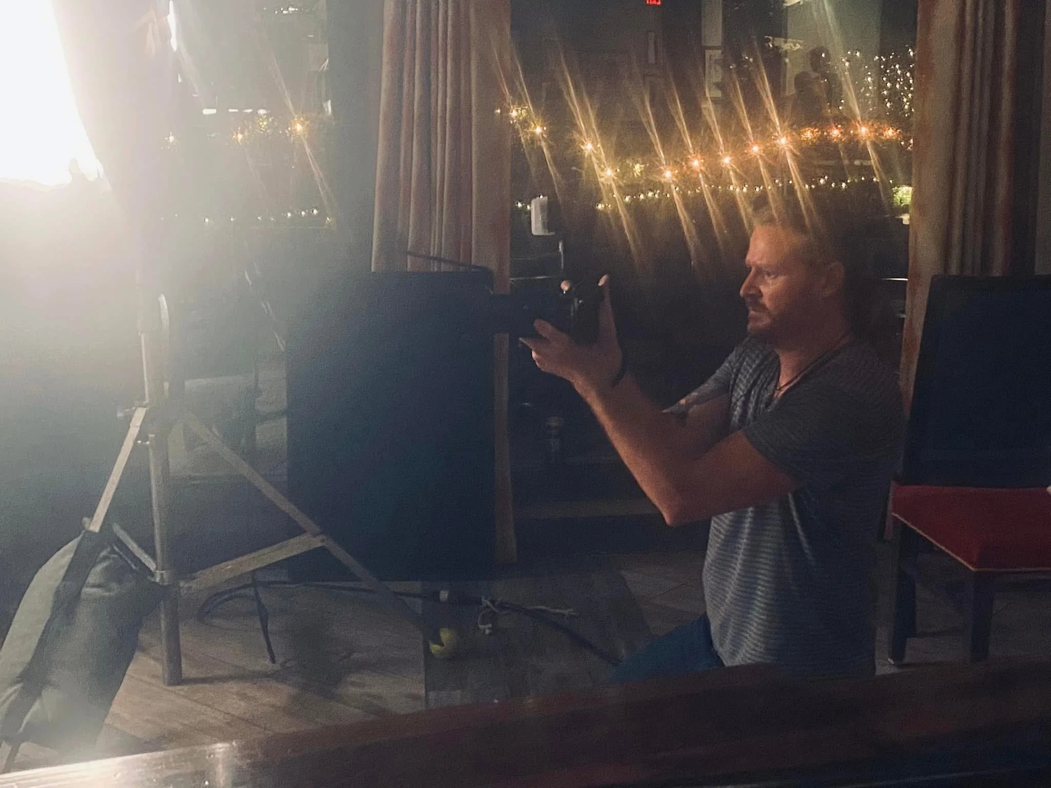 A man with a beard wearing a striped shirt, sitting indoors at night, holding a mobile phone, with a dark curtain and a window with city lights visible behind him.