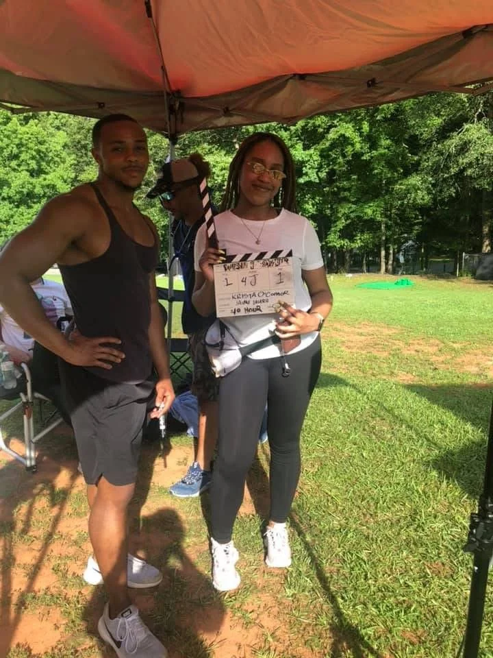 A woman holding a clapperboard stands next to a man under a canopy tent outdoors on grass, with trees in the background. The woman is smiling, wearing glasses, a white t-shirt, and black pants. The man is standing with his hand on his hip, wearing a 