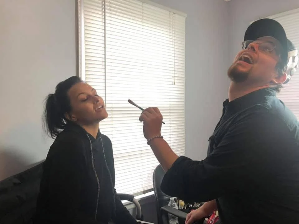 A woman has her eyes closed and is tilting her head as a man applies makeup to her face with a brush.