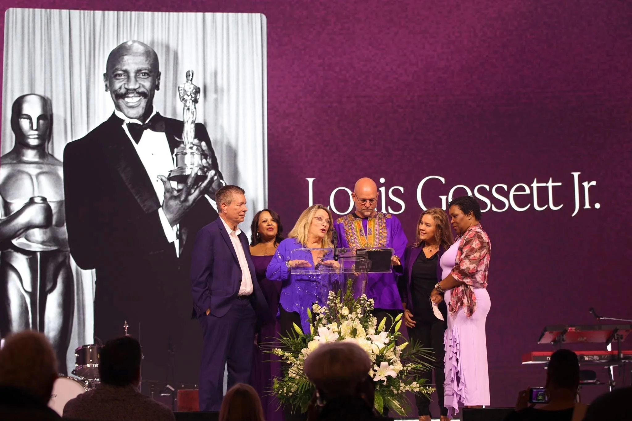 A group of six people on stage at an award ceremony, with a large screen behind them displaying a black and white photo of Louis Gossett Jr. holding an award. The stage is decorated with a floral arrangement. The woman in the purple dress appears to 