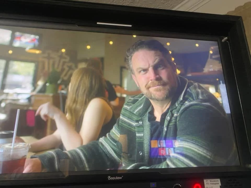 A man with a beard and wavy hair, wearing a striped jacket, sitting in a restaurant or cafe, looking directly at the camera with a serious expression. A woman with long hair sits next to him, her face turned away. There is a drink with a straw in fro