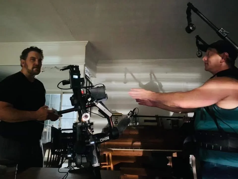 Two men on a film set, one operating a camera and the other gesturing with his hands.