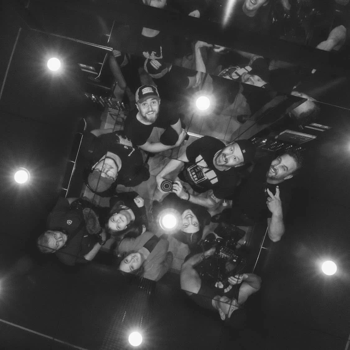 Group of nine friends taking a selfie in a dimly lit room, some smiling and flashing peace signs, with a camera and equipment visible, in black and white.