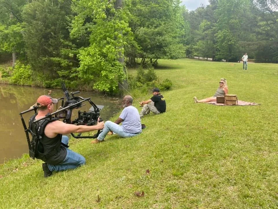 Film crew filming two people fishing by a pond in a park, with a woman relaxing on a blanket nearby.