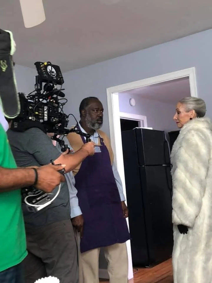 A woman dressed in a white fur coat and black gloves stands facing a man wearing an apron in a doorway. Two men, one holding a camera, are filming the scene inside a home with blue walls.