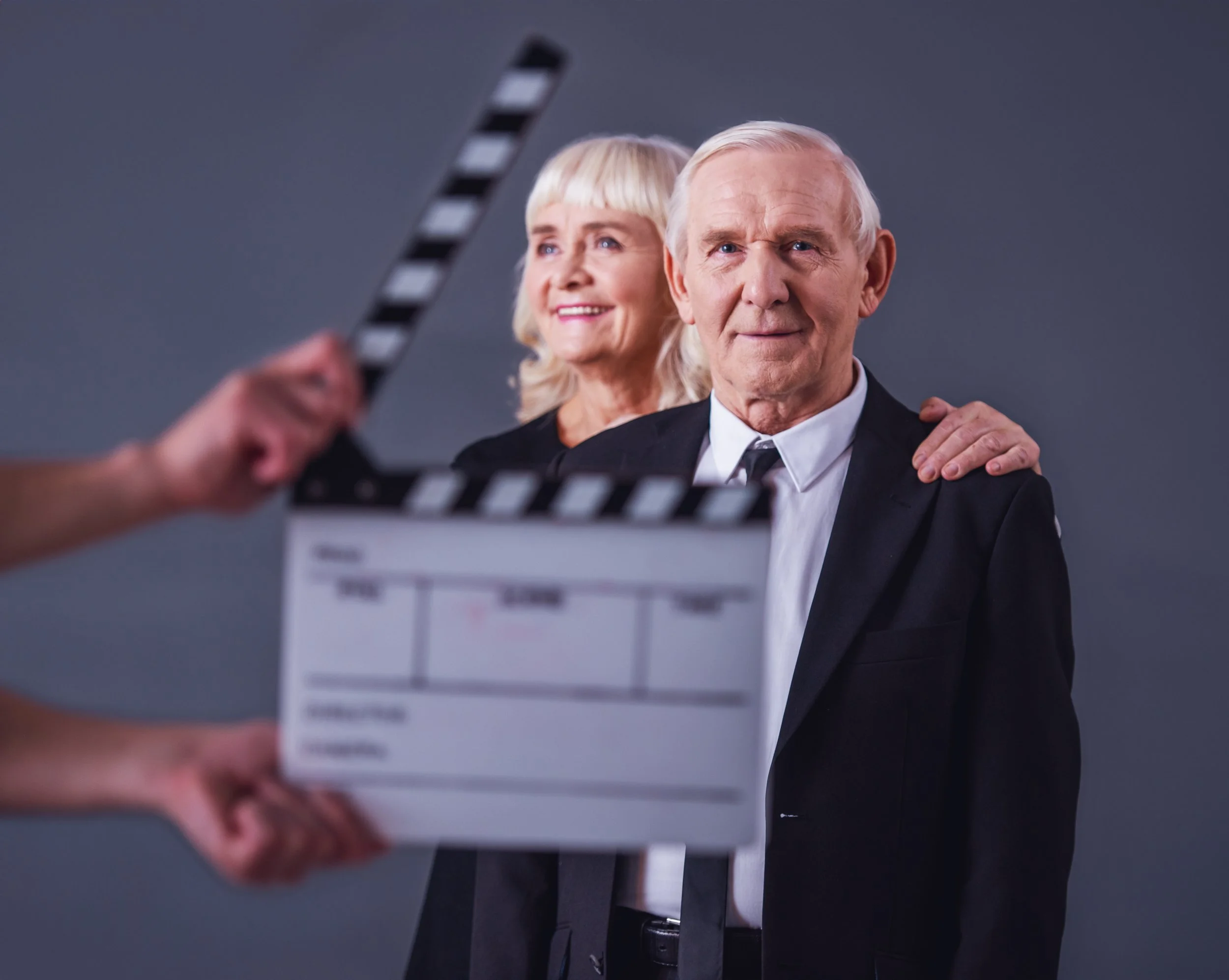 An elderly man and woman in formal attire posing for a photo with a gray backdrop, being filmed or photographed with a clapperboard in the foreground.