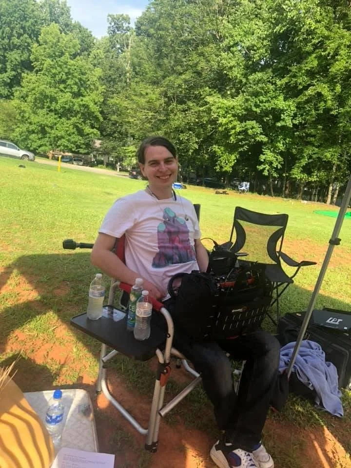 Young man sitting outdoors on a specialized chair with a tray, surrounded by water bottles and equipment, smiling at the camera with a background of green trees and grassy field.