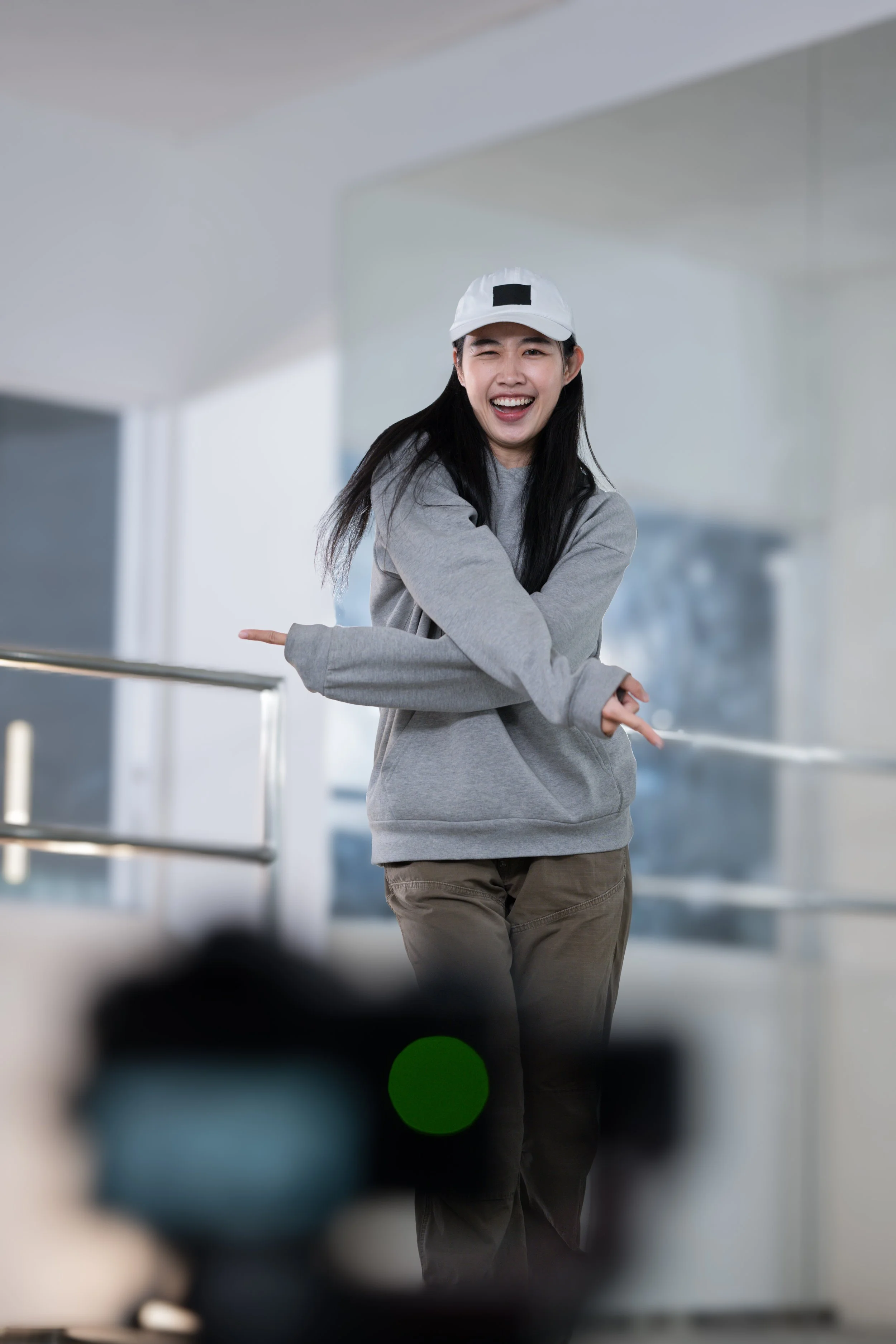 A young woman with long black hair, wearing a white cap with a black square patch, a gray sweatshirt, and khaki pants, is smiling and winking while pointing sideways with both arms in a bright, modern indoor space.