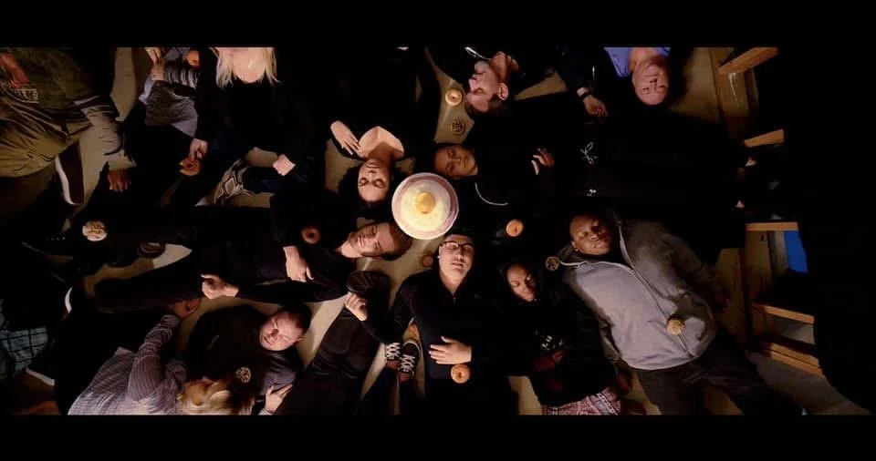 Group of people sitting and standing around a table, viewed from above, with drinks and snacks, in a cozy indoor setting.
