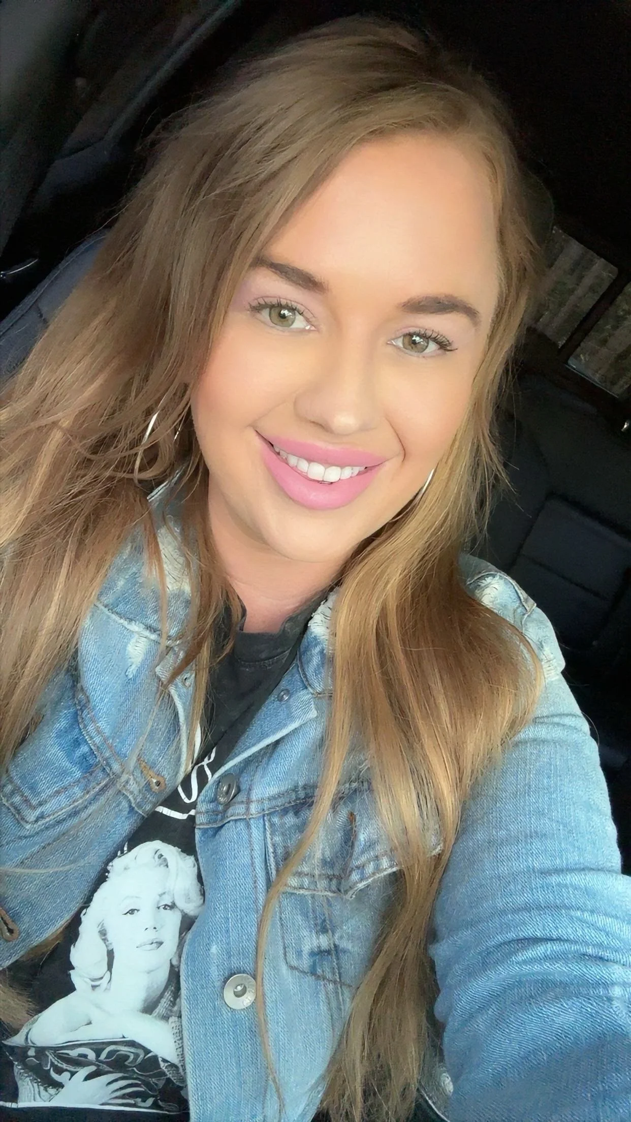 A smiling young woman with long blonde hair, wearing a jean jacket over a Marilyn Monroe t-shirt, sitting inside a vehicle.