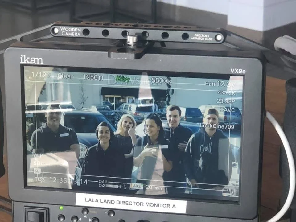 A video camera monitor showing a group of five people smiling and posing for a photo outdoors in a parking lot with cars in the background.