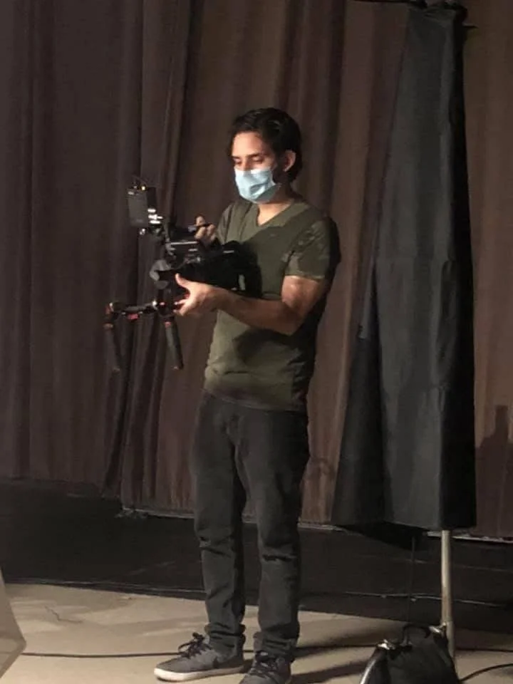 A person wearing a face mask and casual clothing operating a camera on a tripod in front of a dark curtain.